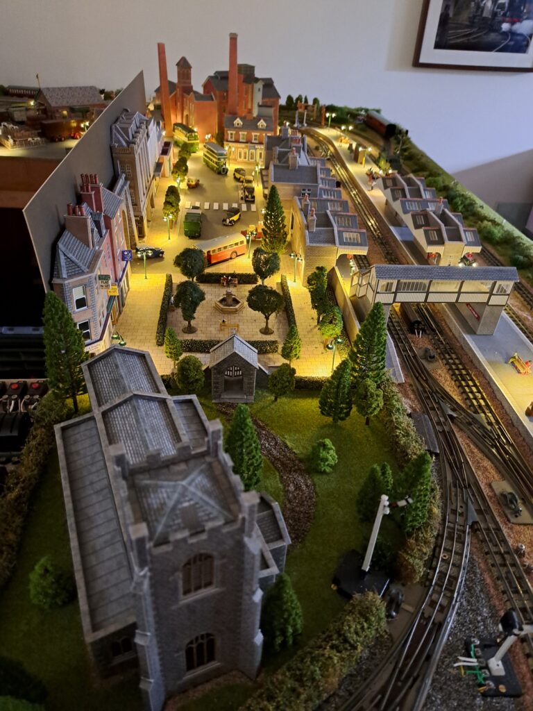 A model railway set-up comprising a night-time scene of a built-up area with tracks and a station. There is a church in the foreground