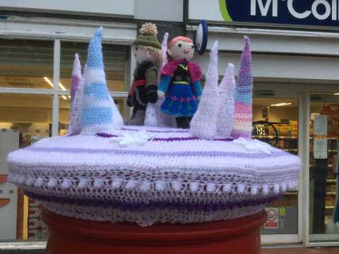 A purple crocheted cover for a post box with figures and conical shapes of ice