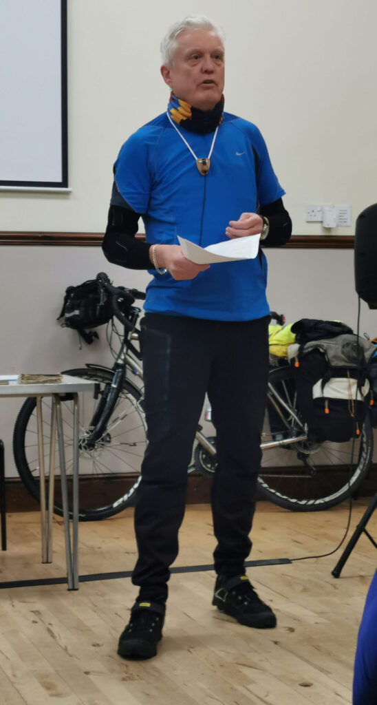 A man in blue top and black trousers standing in front a heavily-laden bike